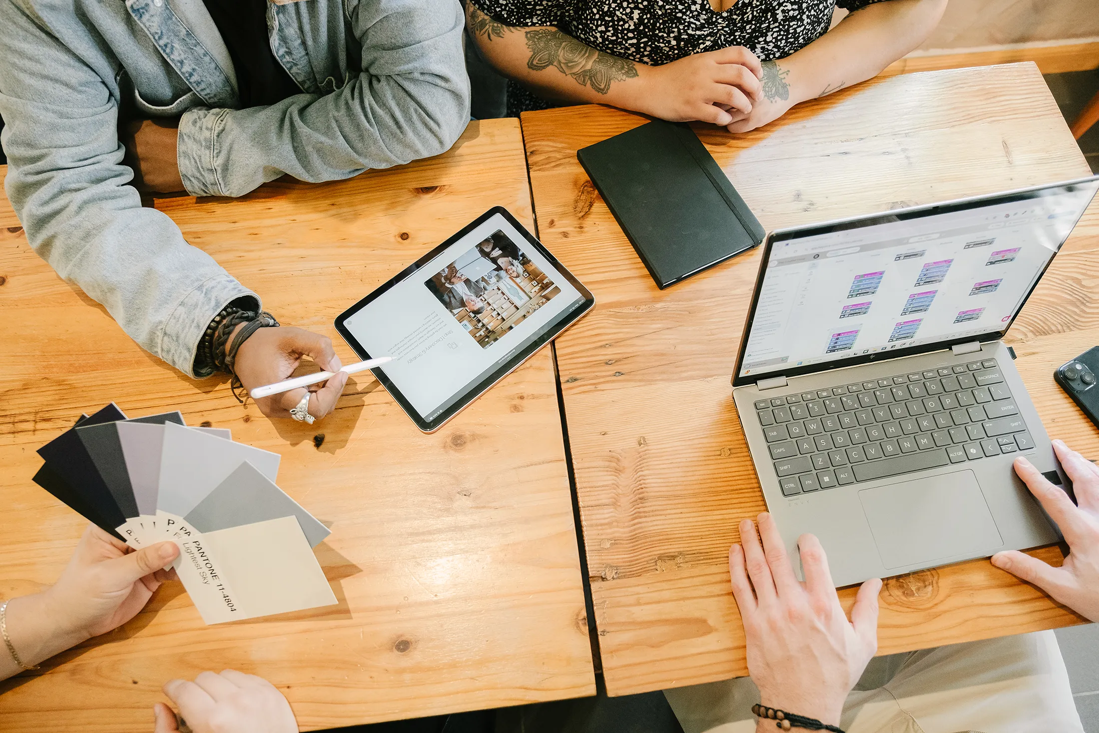 Collaborative Digital Design and Brand Planning Session Team collaborating at a wooden table with a tablet displaying a website layout, a laptop showing content planning, and Pantone color swatches used for brand identity and rebranding work.