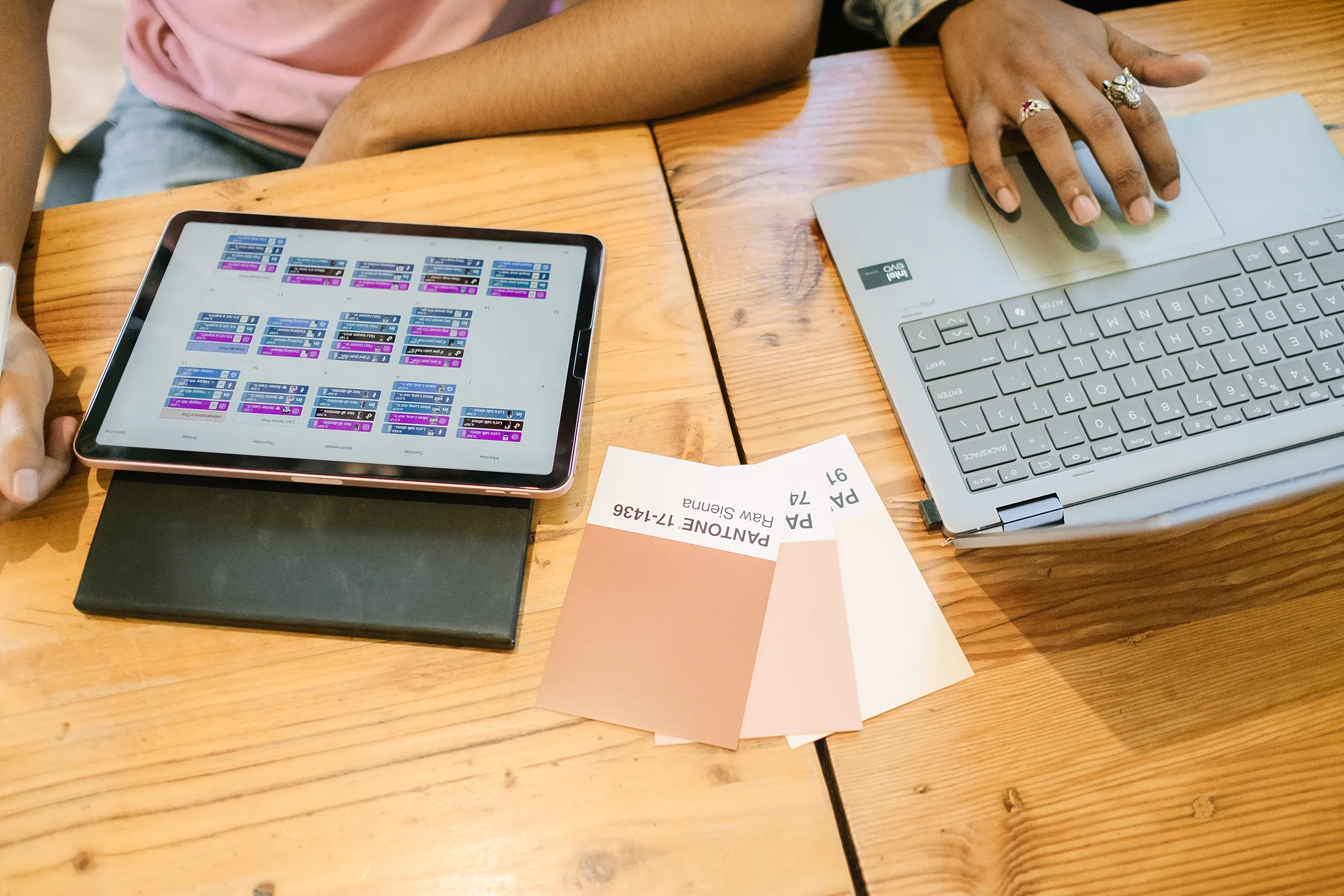 Collaborative Digital Design and Planning Process Two people working at a wooden table with a tablet showing a content schedule, a laptop, and color swatch cards laid out for branding and design planning.